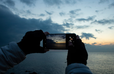 Girl's hand holding smartphone taking sunset photo. Hands in knitted gloves.