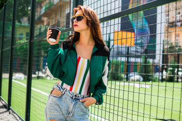 Portrait of a girl in clothes in the style of the 90s, sporty style, jacket, jeans, bananas, sunglasses. Lady on the  play background, drinking morning coffee