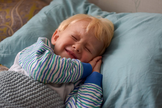 A little blond boy in striped pajamas with his hands under his cheek blinks, trying to sleep