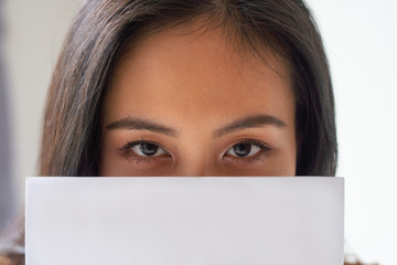 Close-up of Asian lady hiding behind paper and looking at camera. Pretty young woman with document. Businesswoman concept