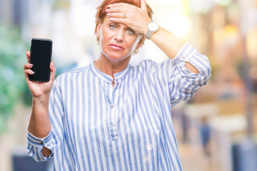 Atrractive senior caucasian redhead woman showing smartphone screen over isolated background stressed with hand on head, shocked with shame and surprise face, angry and frustrated. Fear and upset