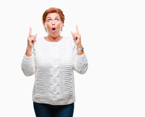 Atrractive senior caucasian redhead woman wearing winter sweater over isolated background amazed and surprised looking up and pointing with fingers and raised arms.