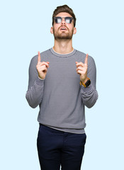 Young handsome man wearing sunglasses amazed and surprised looking up and pointing with fingers and raised arms.