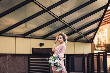 attractive young girl in a short dress with a flower bouquet posing on the street near the building wall.