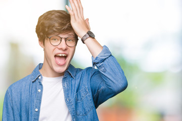 Young handsome man wearing glasses over isolated background surprised with hand on head for...