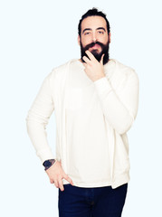 Young man with long hair and beard wearing sporty sweatshirt looking confident at the camera with smile with crossed arms and hand raised on chin. Thinking positive.