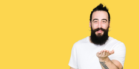 Young hipster man with long hair and beard wearing casual white t-shirt looking at the camera blowing a kiss with hand on air being lovely and sexy. Love expression.