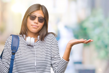 Young asian student woman wearing headphones and backpack over isolated background clueless and confused expression with arms and hands raised. Doubt concept.