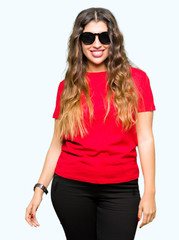 Naklejka premium Young beautiful woman wearing red t-shirt and sunglasses with serious expression on face. Simple and natural looking at the camera.