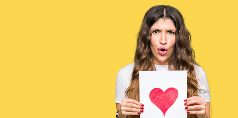 Young adult woman holding card with red heart scared in shock with a surprise face, afraid and excited with fear expression