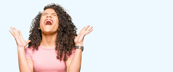 Young beautiful woman with curly hair wearing pink t-shirt celebrating mad and crazy for success...