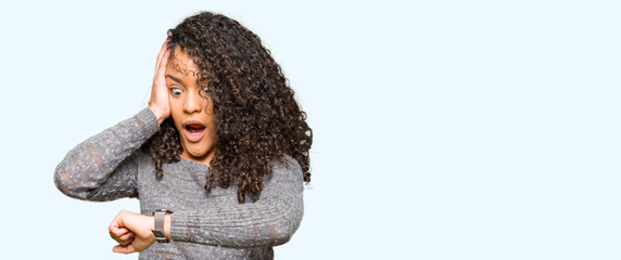 Young beautiful woman with curly hair wearing grey sweater Looking at the watch time worried,...