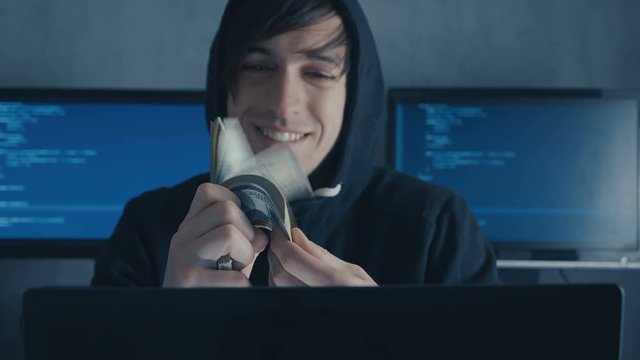 Portrait of Hacker Programmer in hood recounts money earned by hacking. Anonymous hacker recounts dollar bills earned by criminals.