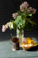 Still life with tea in a silver cup holder with lilac flowers on a black background