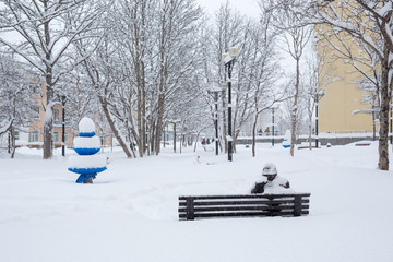 Bench in a snowy city square. After the snowfall there is a lot of snow on the branches of trees and large snowdrifts. Cold winter weather. Feeling of loneliness. Magadan, Siberia, Far East of Russia.