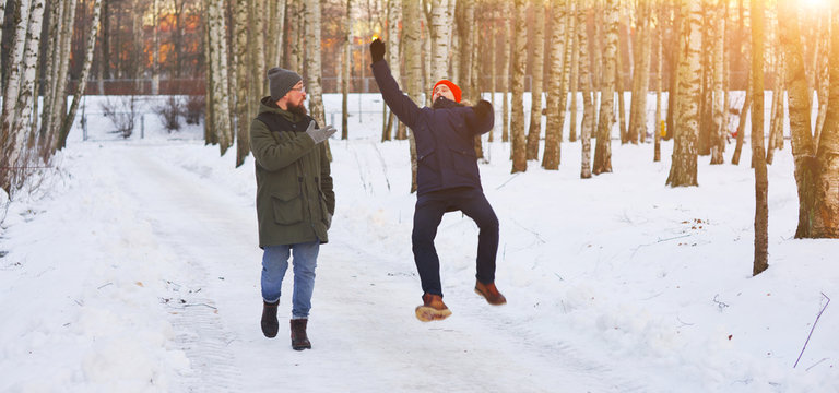 Two Young Modern Man While Walking Through A Birch Forest In Winter. One Of Them Slipped And Lost His Balance, His Friend Held Out His Arms. Freeze Frame Before Falling Into The Snow.