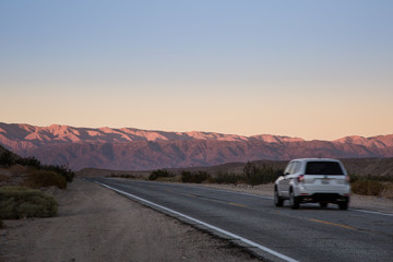 Fototapeta na wymiar White SUV Vehicle Traveling Down an Empty Desert Road at Sunset or Sunrise with Mountains in the Distance