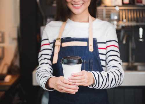 asian woman barista wear jean apron holding hot coffee cup served to customer with smiling face at bar counter,Cafe restaurant service concept.waitress working.