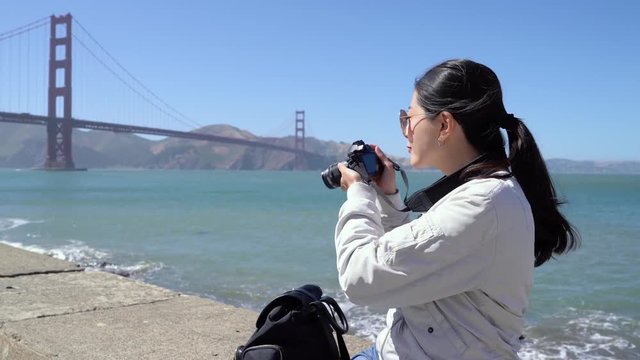 travel tourism and photography concept. happy young asian woman with backpack and camera photographing over golden gate bridge in san francisco sitting on bay on sunny day. girl love photographing.