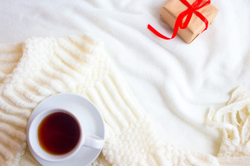 Fototapeta na wymiar A cup with tea or coffee and a box with a gift tied with a red ribbon on a white knitted background. Romantic breakfast in bed for Valentine's Day or a gift for mother's day 