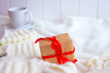 A cup with tea or coffee and a box with a gift tied with a red ribbon on a white knitted background. Romantic breakfast in bed for Valentine's Day or a gift for mother's day
