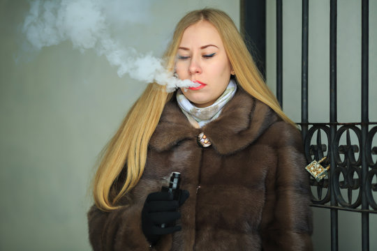 Vape lady. Young pretty girl blonde in a fur coat smokes an electronic cigarette and lets off puffs of steam near vintage gateway outdoors in the winter.