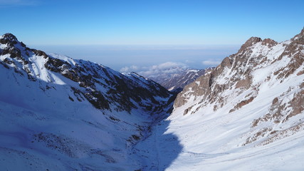 View from the snowy gorge in the mountains to the clouds and the city smog. Coniferous trees and firs are visible. Snowy mountains and grey ground. Gradient blue sky and dark clouds. Drone.