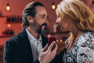 A middle-aged man and woman look into each other’s eyes. A man in one hand holds a box with a wedding ring.