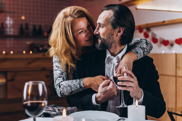 Man raises a glass of wine while a woman hugs him from behind in a restaurant