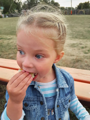 a little girl of Caucasian appearance with blond hair and blue eyes in denim clothes eating a bun in the Park.