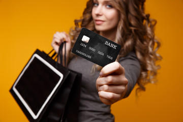 lose-up portrait of happy young brunette woman holding credit card and black shopping bags, looking at camera, isolated on yellow background
