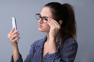 Woman with bad sight trying to read message on screen of mobile phone against grey background