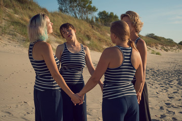 Four women stand in a circle and hold hands on the beach