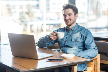 Portrait of handsome happy bearded young businessman in blue jeans shirt are sitting in cafe and have a break with cup of coffee and toothy smile are looking to the laptop.