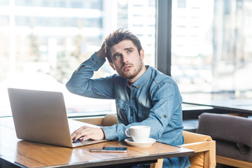 Fototapeta na wymiar Portrait of handsome thoughful bearded young businessman in blue jeans shirt are sitting in cafe and typing message on laptop, having new idea and planning own strategy, holding one hand on the head.
