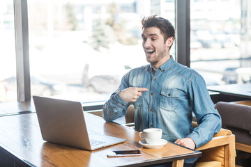Fototapeta na wymiar Portrait of happy toothy smiling bearded young freelancer in blue jeans shirt are sitting alone in cafe and having new proposal for a work on laptop are speaking for webcame in positive mood.