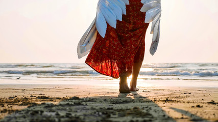 Beautiful female angel walking barefoot toward the sea at sunset.