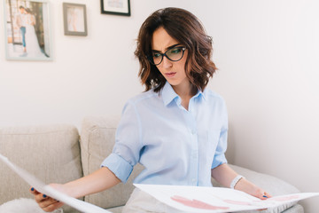 It is a portrait of a brunette young girl sitting on the sofa in studio. She wears blue shirt and white watch . She is curious about sketches which she holds in both hands.