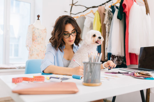 A brunette girl in a blue shirt is sitting at the table in the workshop . She is deawing some sketches. The dog is sitting on the knees.
