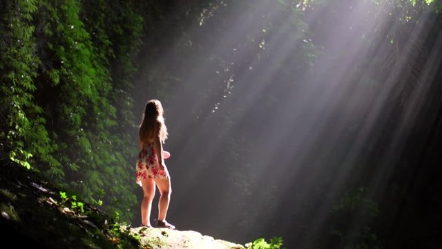 Girl walks into sun rays and reaches out with hand at beautiful Tukad Cepung Waterfall. Slow motion.