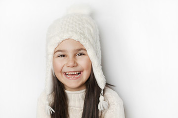Happy cute child girl in a white winter hat and a white knitted sweater laughs on a white background. Real positive emotions. Smiling beautiful face. 