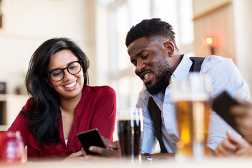 leisure, technology and people concept - happy man and woman with smartphones and drinks at bar or restaurant