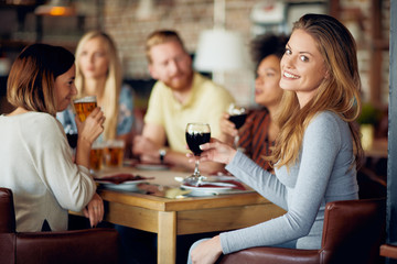 Woman looking at camera and holding glass of wine while sitting in restaurant. In background friends drinking and chatting.