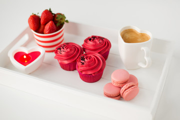 valentines day and sweets concept - close up of cupcakes with red buttercream frosting, macarons, heart shaped coffee cup, candle and strawberries on tray