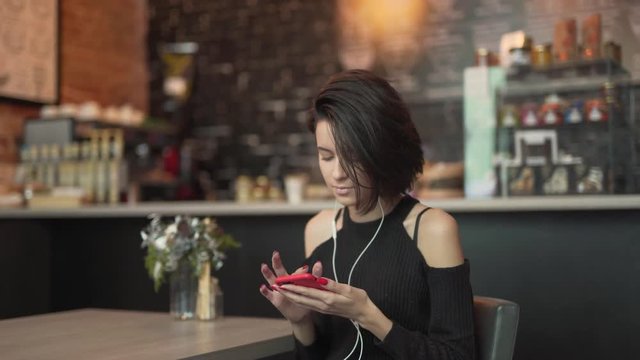 Girl is sitting at the caf&radic;&copy;. She is listening to music in headphones and hold phone in hand. With other hand she fix her short hair.