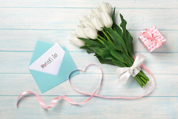 A bouquet of white tulips and a pink ribbon in the form of a heart with a gift box, love note and color envelope on blue wooden boards. Mother's day