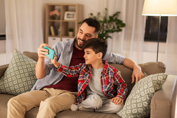 people, family and technology concept - happy father and little son taking selfie by smartphone sitting on sofa at home
