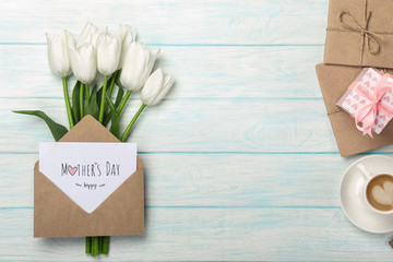 A bouquet of white tulips, cup of coffee, gift box with a love note and envelope on blue wooden boards . Mother's day