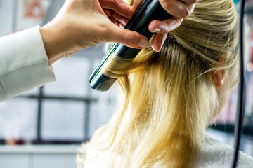 Fototapeta premium Close-up of hairdresser holding strand of girls blonde hair with flat iron making beautiful curls