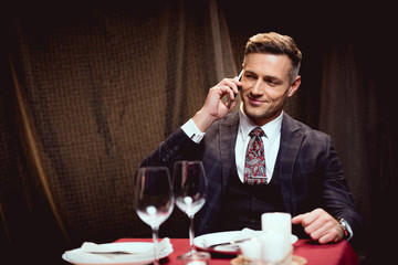 handsome smiling man in suit sitting at table and talking on smartphone in restaurant